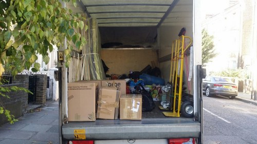 The image shows the open rear of a white commercial van parked on a residential street, with the trunk area filled with a variety of waste materials ready for removal. In the foreground, several cardboard boxes of different sizes and shapes are stacked, some sealed with tape and others partially open, revealing contents such as plastic packaging and paper waste. To the right, a yellow hand trolley or dolly is leaning against the van's interior wall, accompanied by a small black and silver wheeled bin or container. Behind the boxes, there are miscellaneous discarded items including plastic bags, packaging materials, and some loose rubbish, all indicating commercial waste ready for collection. The surroundings include a paved pavement with a curb, a small tree with green leaves on the left, and a residential street with parked cars and houses visible in the background, suggesting a suburban area. The lighting appears natural, with daylight illuminating the scene, consistent with typical waste collection operations in Ruislip or nearby areas.