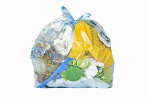 A clear, transparent plastic rubbish bag filled with various waste items, including yellow plastic packaging, green plastic bottle caps, and pieces of paper and plastic debris, is placed on a plain white background. The bag appears to be loosely packed but generally maintains its shape, with the top gathered and tied off. The contents inside are visibly mixed, with some items pressing against the plastic, creating uneven surfaces. The waste materials suggest typical household or small commercial refuse, consisting of lightweight plastics, paper waste, and plastic bottle caps. The smooth surface and semi-glossy finish of the plastic bag reflect light, highlighting the different textures and colors of the waste within. This type of waste collection is often managed by rubbish removal services such as Commercial Waste Removal Ruislip, who handle disposal and clearance of general waste collected from businesses or domestic properties in local areas like Ruislip, adjacent to London. The image emphasizes the importance of proper waste containment before collection or disposal as part of responsible rubbish management practices.