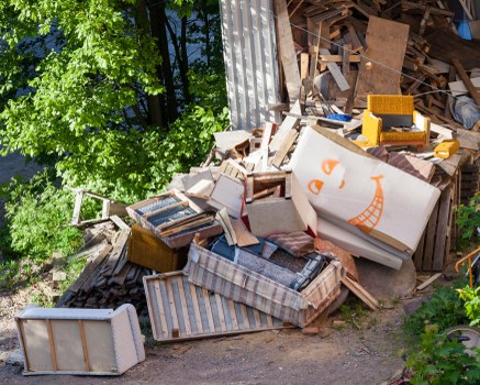 A large pile of discarded furniture and wooden debris situated outdoors, with a mix of broken and intact items including a striped mattress, wooden cabinets, and a cardboard box with an orange smiley face. The furniture shows varying textures, from smooth painted surfaces to rough, weathered wood. The debris is stacked haphazardly on a paved or concrete area adjacent to a tall metal fence, partly shaded by a leafy tree with bright green foliage visible in the background. Some items appear to be discarded company or household waste, and the scene likely depicts a collection site for rubbish removal services in Ruislip, as offered by Commercial Waste Removal Ruislip, with natural sunlight illuminating the clutter and foliage around it.
