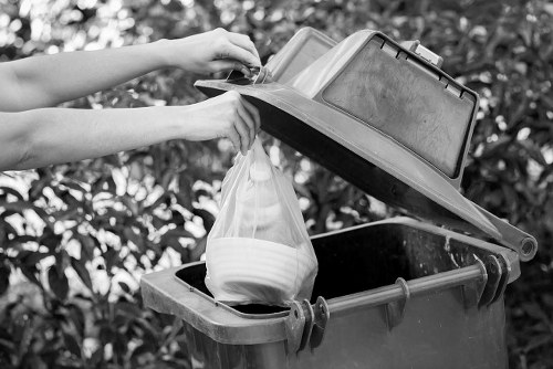 A person’s arms are seen lifting the lid of a dark grey or black outdoor rubbish bin, located on a paved driveway or street, with a background of dense, leafy foliage. The open bin reveals a white plastic bag filled with mixed waste, partially protruding from the top. The bin’s lid is hinged, made of sturdy plastic, and appears weather-resistant. The scene suggests a typical waste collection area, potentially in a residential or semi-urban setting near Ruislip, with natural light illuminating the area. This visual context aligns with professional rubbish removal and waste management services provided by Commercial Waste Removal Ruislip and illustrates typical waste disposal tasks done in the local area.