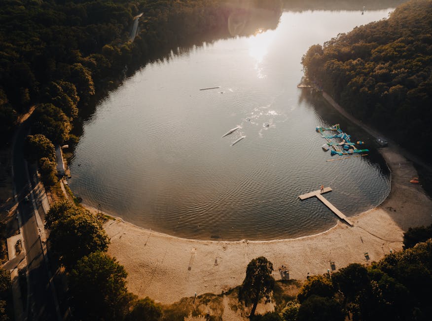 An aerial view of a small, semi-circular sandy beach at the edge of a calm lake surrounded by dense, green forested hills. The lake's water is still and reflects the sky with a bright, diffused sunlig
