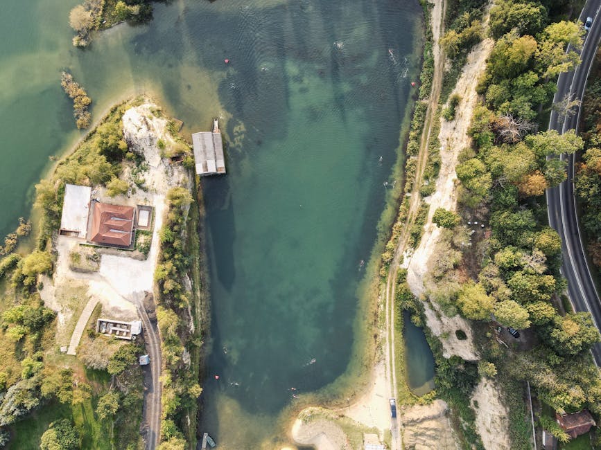 An aerial view of a small, semi-circular sandy beach at the edge of a calm lake surrounded by dense, green forested hills. The lake's water is still and reflects the sky with a bright, diffused sunlig