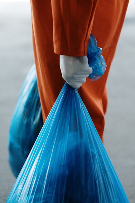 A sanitation worker dressed in a blue uniform with an orange high-visibility vest is positioning a blue wheeled bin next to a garbage collection truck on a narrow city street. The truck, predominantly